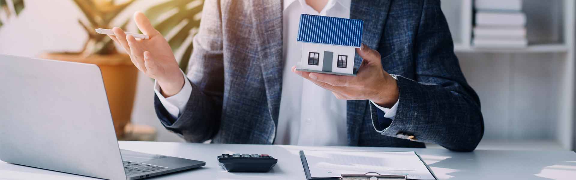 A real estate professional in a gray blazer holding a small house model while sitting at a desk with a laptop, calculator, and clipboard.
