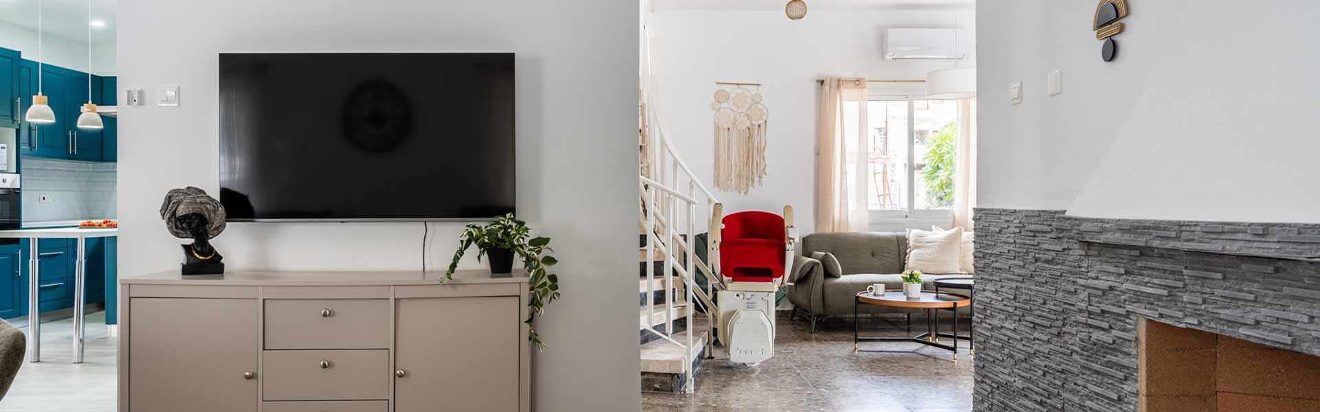 The bright and modern Deyar house interior featuring a living area with a red stairlift installed on the staircase, a large wall-mounted TV, and a glimpse of a teal-colored kitchen.